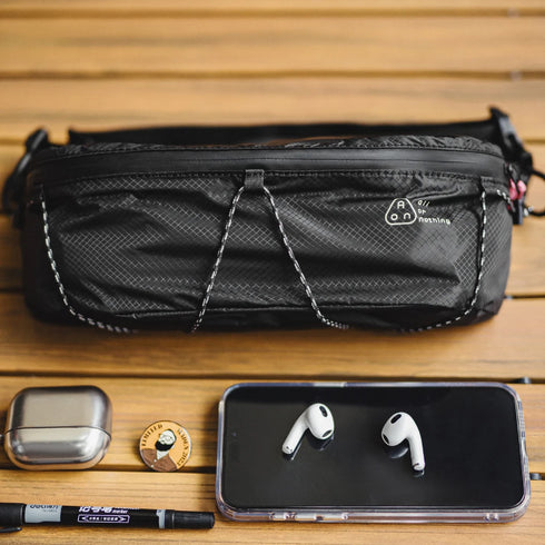 Black waist bag with phone, earbuds, and pen on a wooden surface